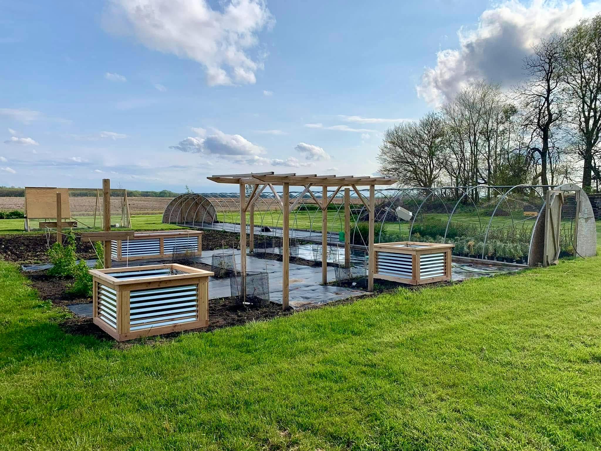 Wide view of a working production garden — cedar and galvanized raised beds in front, pergola in middle, hoop houses behind, open field stretching to horizon